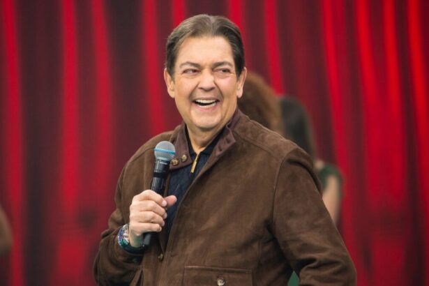 Fausto Silva sorrindo, de camisa marrom, segurando o microfone com a mão direita durante gravação do seu programa na Band