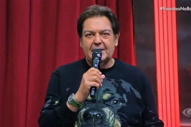 Fausto Silva com microfone na mão direita, de camisa preta estampada com um cachorro durante apresentação do Faustão na Band