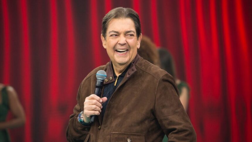 Fausto Silva sorrindo, de camisa marrom, segurando o microfone com a mão direita durante gravação do seu programa na Band