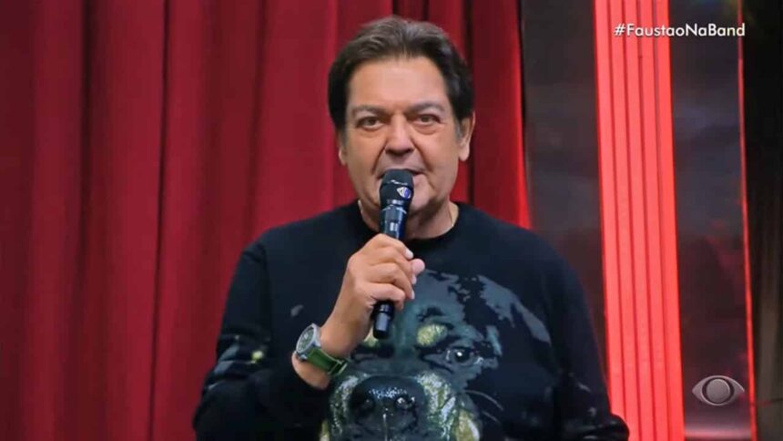 Fausto Silva com microfone na mão direita, de camisa preta estampada com um cachorro durante apresentação do Faustão na Band