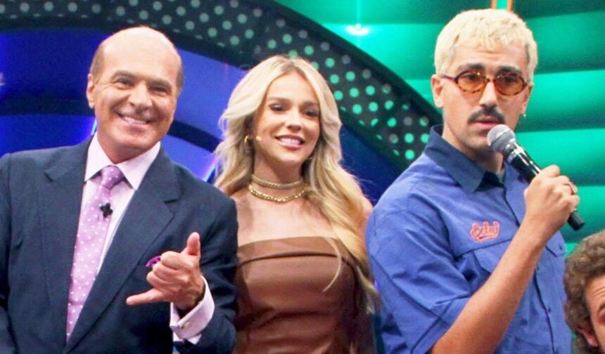 Marcelo de Carvalho com palitó azul, camisa roxa claro e gravata fazendo um hangloose, ao lado de Gabi Lopes, sorrindo, com colar e vestido marrom; vocalista de Jovem Dionísio com figurino azul, oculos e microfone na mão