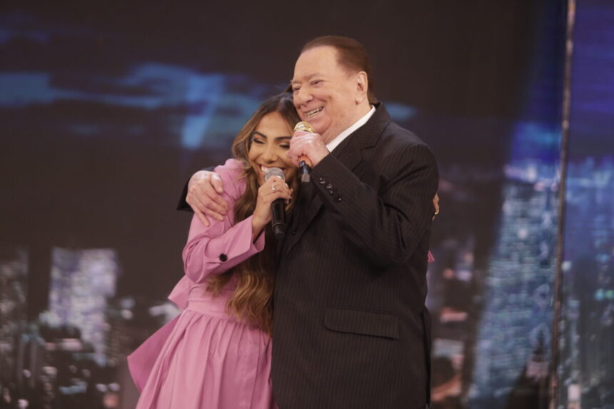 Gabriela Rocha de vestido roxo, sorrindo, abraçando Raul Gil, que está de paltió, gratava e calça preta, sorrindo durante gravação do quadro Homenagens do Programa Raul Gil, do SBT