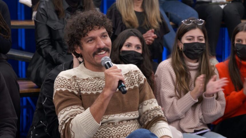 Guito, que interpreta Tibério em "Pantanal", de camisa manga longa marron, bege e branco, segurando o microfone com a mão direita e sorrindo durante entrevista do Altas Horas, da TV Globo