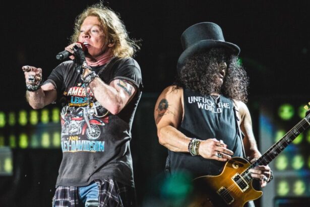 Vocalista Axl Rose cantando, de camisa preta estampada, de luva e calça jeans, com o microfone na mão esquerda; Guitarrista Slash tocando instrumento, de chapéu, camisa regata e calça pretas.