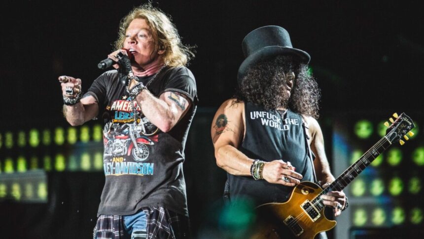 Vocalista Axl Rose cantando, de camisa preta estampada, de luva e calça jeans, com o microfone na mão esquerda; Guitarrista Slash tocando instrumento, de chapéu, camisa regata e calça pretas.