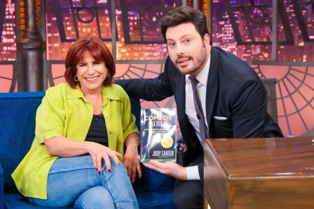 Judy Carter sorrindo, sentada com as pernas cruzadas e mão direita no joelho direito, de camisa verde e preta e calça jeans ao lado de Danilo Gentili, com seu livro em mãos, em sua bancada no The Noite, do SBT