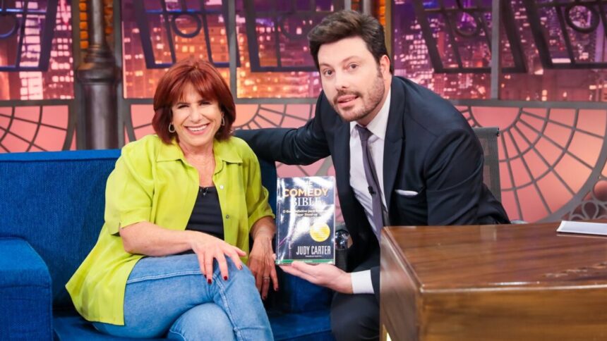 Judy Carter sorrindo, sentada com as pernas cruzadas e mão direita no joelho direito, de camisa verde e preta e calça jeans ao lado de Danilo Gentili, com seu livro em mãos, em sua bancada no The Noite, do SBT