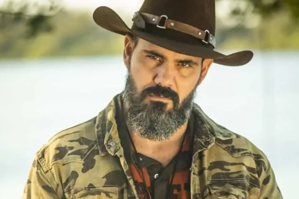 Juliano Cazarré com camisa camuflada, sério, de chapéu, durante gravação de pantanal