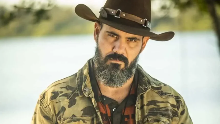 Juliano Cazarré com camisa camuflada, sério, de chapéu, durante gravação de pantanal