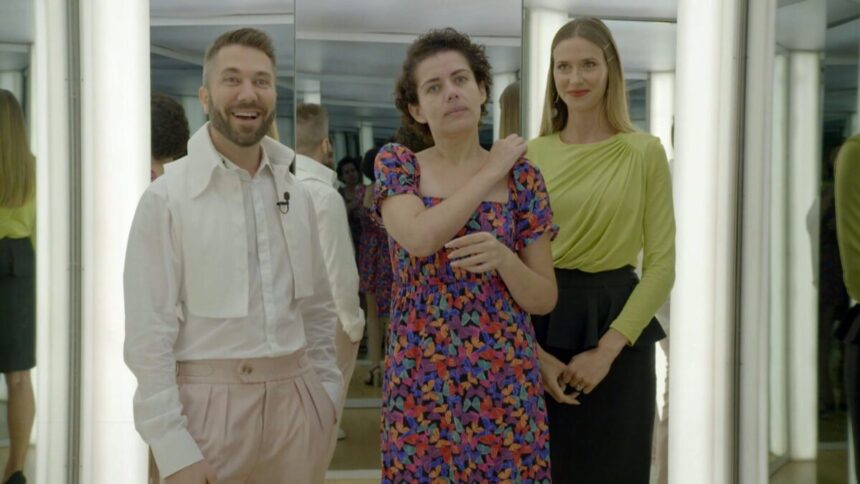 Karina com a mão no ombro, ajeitando um dos vestidos que ganhou da produção; ao lado dos estilistas Lucas Anderi, sorrindo, com a mão no bolso, calça bege e camisa social branca; e Renata Kuerten sorrindo, de camisa manga longa verde e saia preta durante gravação do Esquadrão da Moda, do SBT