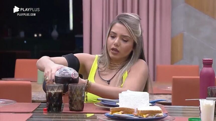 Karol sentada na mesa com uma garrafa de refrigerante nas mãos.