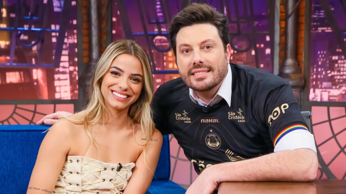 Key Alves de vestido bege, sorrindo, com as mãos no joelho, sentada num sofá azul ao lado de Danilo Gentili, de camisa preta do time de Key, sorrindo, com a mão na bancada e a mão no ombro da convidada, no The noite, do SBT