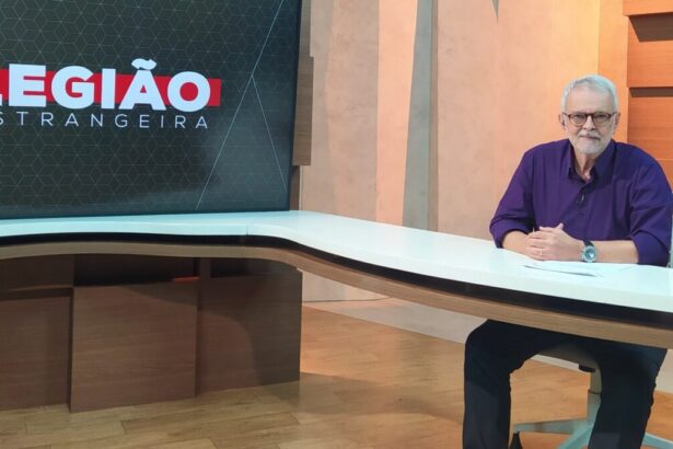 Alberto Gaspar sentado em sua bancada, de camisa azul e calça preta, sorrindo, durante gravação do Legião Estrangeira, da TV Cultura