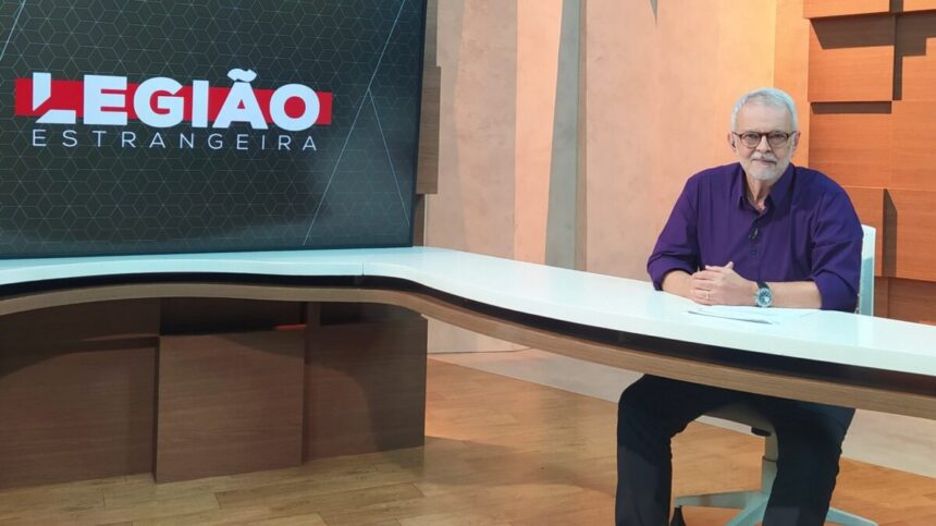 Alberto Gaspar sentado em sua bancada, de camisa azul e calça preta, sorrindo, durante gravação do Legião Estrangeira, da TV Cultura