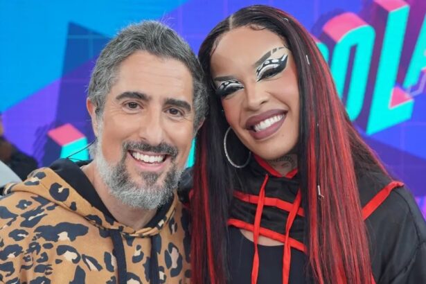 Marcos Mion sorrindo, de camisa animal-print ao lado de Gloria Groove com look e peruca vermelha durante gravação do Caldeirão, da TV Globo