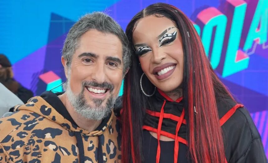 Marcos Mion sorrindo, de camisa animal-print ao lado de Gloria Groove com look e peruca vermelha durante gravação do Caldeirão, da TV Globo