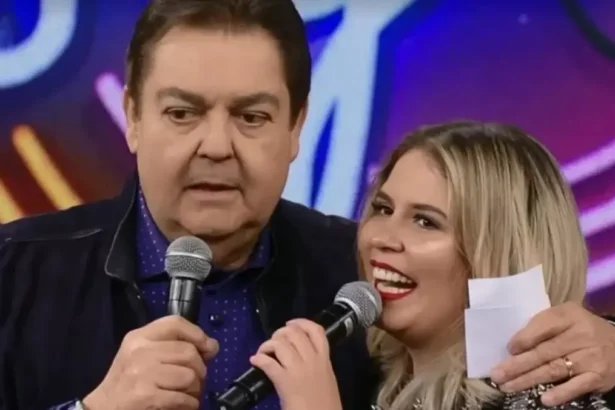 Faustão de camisa preta, segurando o microfone com a mão direita, abraçando Marília, que está de vestido preto, sorrindo, com o microfone próximo a boca, durante gravação do Domingão do Faustão, da TV Globo