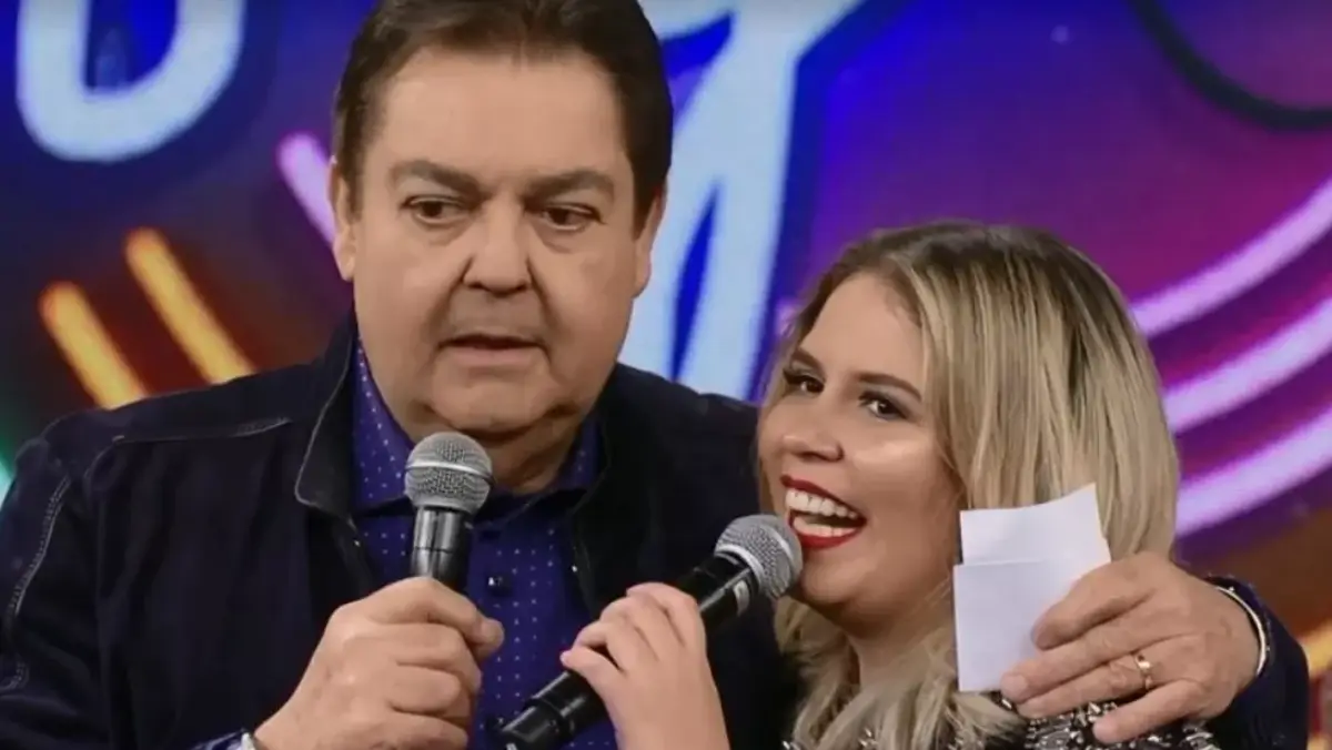 Faustão de camisa preta, segurando o microfone com a mão direita, abraçando Marília, que está de vestido preto, sorrindo, com o microfone próximo a boca, durante gravação do Domingão do Faustão, da TV Globo