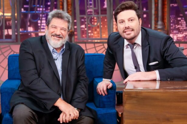 Mario Sergio de palitó preto, mãos juntas do joelho, sentado, sorrindo, ao lado de Danilo Gentili, de palitó, gravata e calça preta, sorrindo, com a mão no sofá azul e outra na bancada do programa, durante gravação no SBT