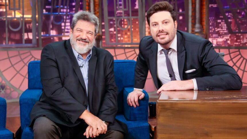 Mario Sergio de palitó preto, mãos juntas do joelho, sentado, sorrindo, ao lado de Danilo Gentili, de palitó, gravata e calça preta, sorrindo, com a mão no sofá azul e outra na bancada do programa, durante gravação no SBT