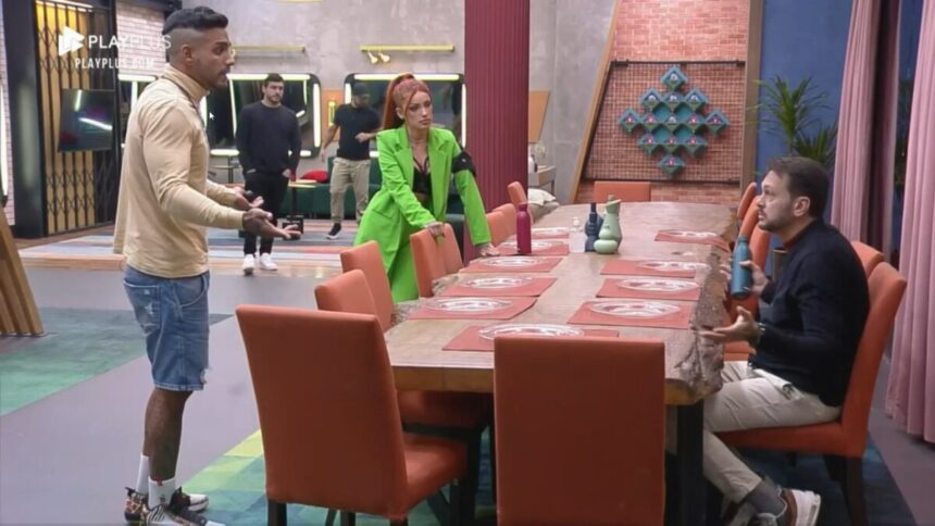 Albert sentado na mesa do Power Couple Brasil discutindo com Matheus e Brenda que estão em pé