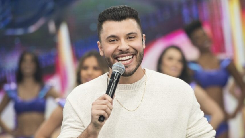 Murilo Huff com camisa manga-longa branca, sorrindo, com microfone na mão esquerda durante apresentação no Faustão na Band