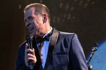 Leandro Hassum de palitó e gravata de laço pretos, camisa branca, com microfone na mão esquerda durante gravação do filme O Auto da Boa Mentira