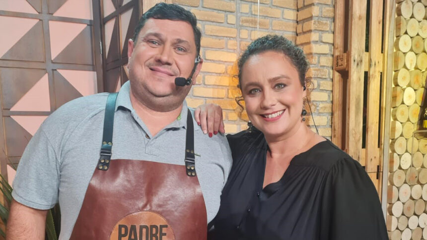 Padre Chef Evandro de camisa social cinza e avental marrom com a logo do programa Sabor de Vida e Bianca Lauá de camisa social preta, colar de ouro, sorrindo, com a mão no ombro do Chef durante gravação de Sabor de Vida, da TV Aparecida