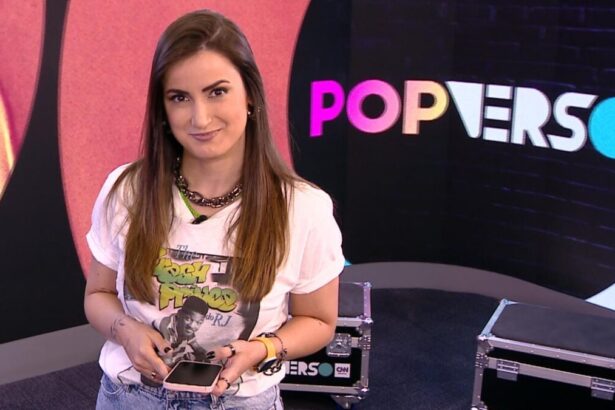 Mari Palma sorrindo, de camisa branca estampada, segurando um celular com as mãos durante gravação do Popverso, da CNN Brasil