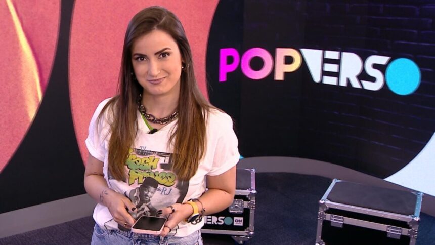 Mari Palma sorrindo, de camisa branca estampada, segurando um celular com as mãos durante gravação do Popverso, da CNN Brasil