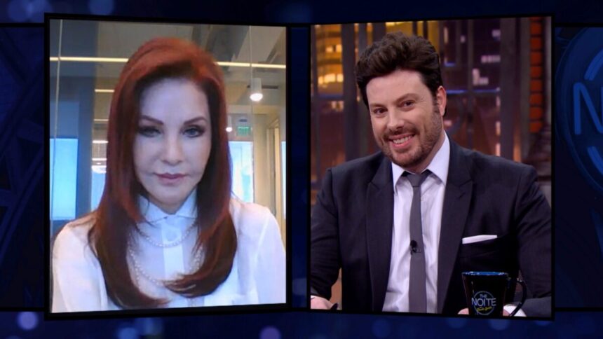 Priscila Presley de camisa branca, cabelo solto, sentada; Danilo Gentili de palitó, gravata e camisa social branca, sentado, sorrindo, na sua bandaca, durante gravação de entrevista