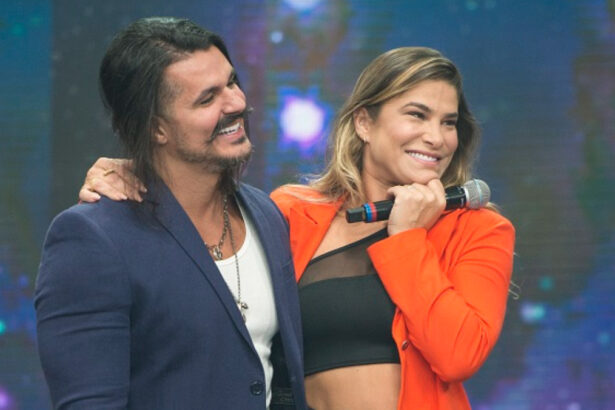 Bruno Lopes de blazer azul, camisa branca e calça jeans, abraçando Priscila Fantin, que está de blazer vermelho e top preto, sorrindo, com microfone na mão, durante gravação do Faustão na Band