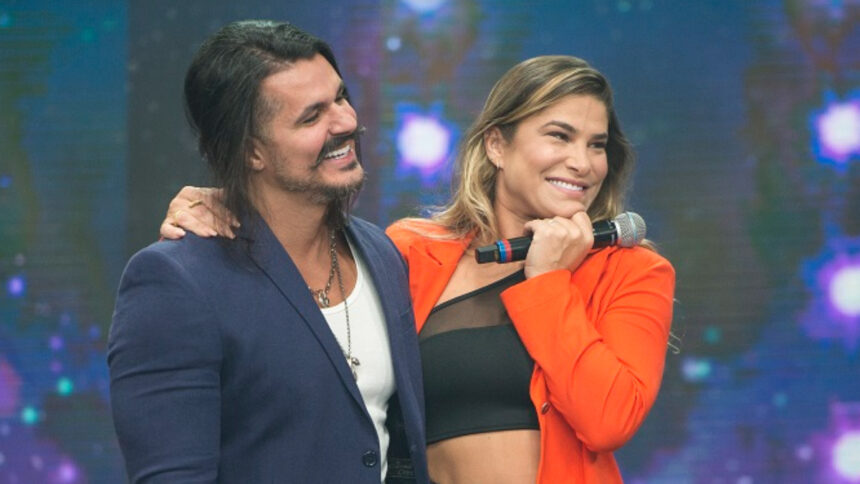 Bruno Lopes de blazer azul, camisa branca e calça jeans, abraçando Priscila Fantin, que está de blazer vermelho e top preto, sorrindo, com microfone na mão, durante gravação do Faustão na Band