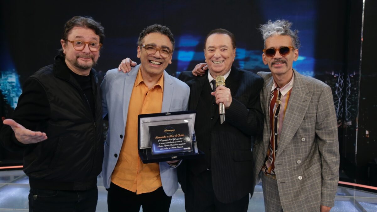 Raul Gil de palitó e gravata preta, microfone dourado na mão esquerda, sorrindo, ao lado de Simoninha, segurando uma placa de homenagem ao pai, Wilson Simonal