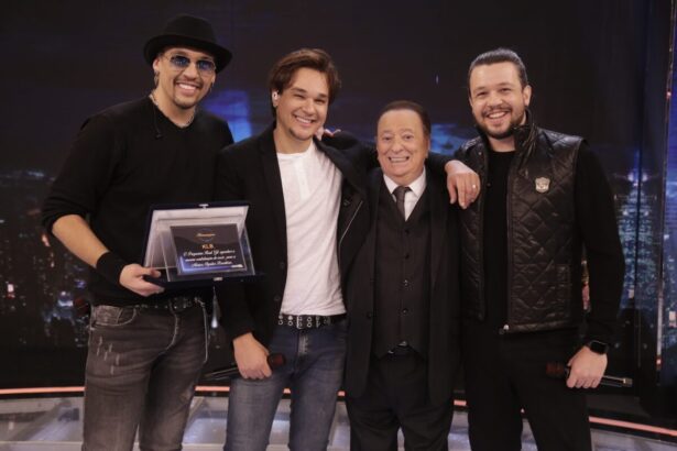 Kiko segurando a placa de homenagem ao lado de Leandro Raul Gil e Bruno, ambos preto e branco, sorrindo, durante gravação do Programa Raul Gil, do SBT