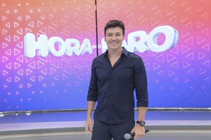 Rodrigo Faro sorrindo em frente ao logo do seu programa, no cenário da Record TV