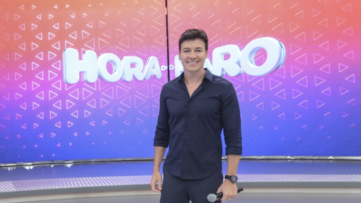 Rodrigo Faro sorrindo em frente ao logo do seu programa, no cenário da Record TV