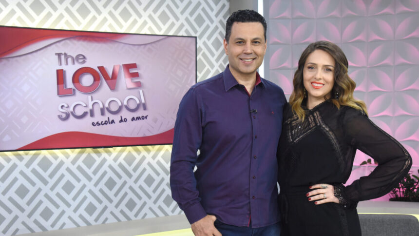 Renato e Cristiane Cardoso abraçados nos estúdios do The Love School, com a televisão ao fundo tendo o logo do programa