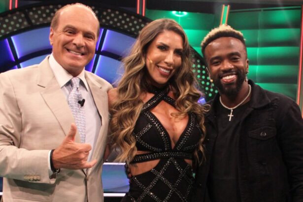 Tati Minerato com vestido decotado preto brilhoso, sorrindo com a mão no ombro de Negrete, que está sorrindo, de colar de ouro e camisa preta; Marcelo de Carvalho sorrir, fazendo hangloose com a mão direita, usando blazer cinza e gravata azul clara durante gravação do seu programa na RedeTV!