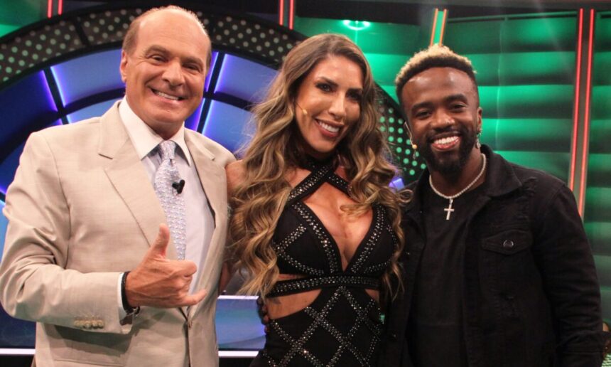 Tati Minerato com vestido decotado preto brilhoso, sorrindo com a mão no ombro de Negrete, que está sorrindo, de colar de ouro e camisa preta; Marcelo de Carvalho sorrir, fazendo hangloose com a mão direita, usando blazer cinza e gravata azul clara durante gravação do seu programa na RedeTV!