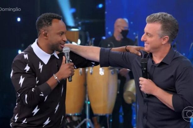 Thiaguinho com camisa manga longa preta e branco com a mão no ombro de Luciano Huck, sorrindo, com o microfone preto na mão direita; Luciano Huck com camisa social preta, sorrindo, com microfone preto na mão esquerda com a mão no ombro de Thiaguinho durante gravação do Domingão com Huck, na TV Globo