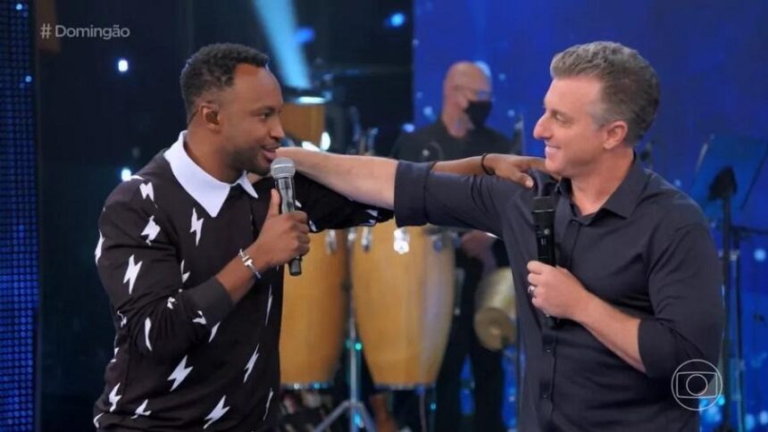 Thiaguinho com camisa manga longa preta e branco com a mão no ombro de Luciano Huck, sorrindo, com o microfone preto na mão direita; Luciano Huck com camisa social preta, sorrindo, com microfone preto na mão esquerda com a mão no ombro de Thiaguinho durante gravação do Domingão com Huck, na TV Globo