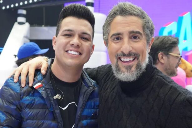 Vitor Fernandes sorrindo de casaco preto com azul, abraçando Marcos Mion, que está de camisa manga-longa preta, sorrindo, durante gravação do Caldeirão, da TV Globo