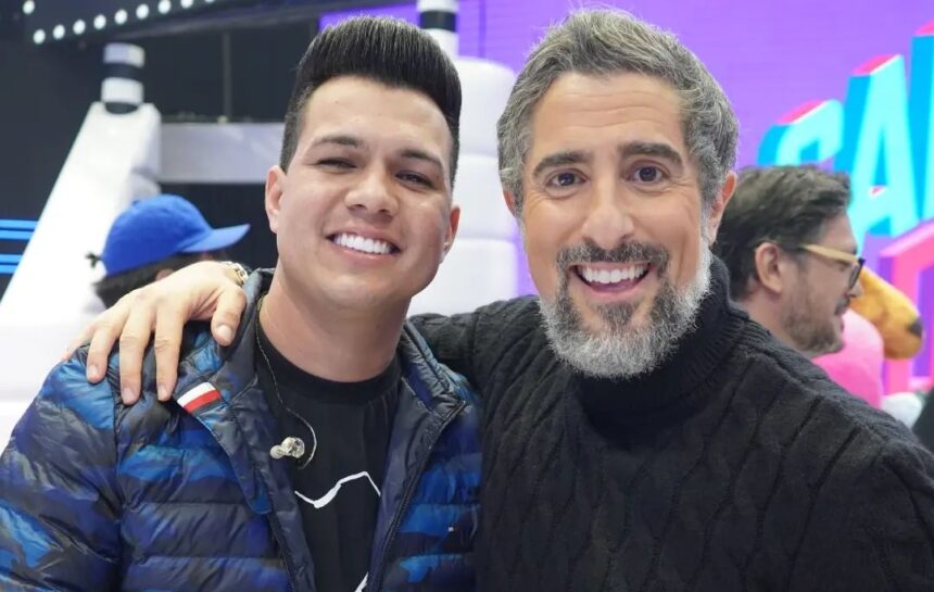 Vitor Fernandes sorrindo de casaco preto com azul, abraçando Marcos Mion, que está de camisa manga-longa preta, sorrindo, durante gravação do Caldeirão, da TV Globo