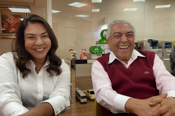 Mônica sorrindo, de camisa branca, sentada, de cabelo solto ao lado de Mauricio de Sousa, seu pai, de camisa social branca e sobreposição vinho, sorrindo durante gravação do Conversa com Bial, da TV Globo