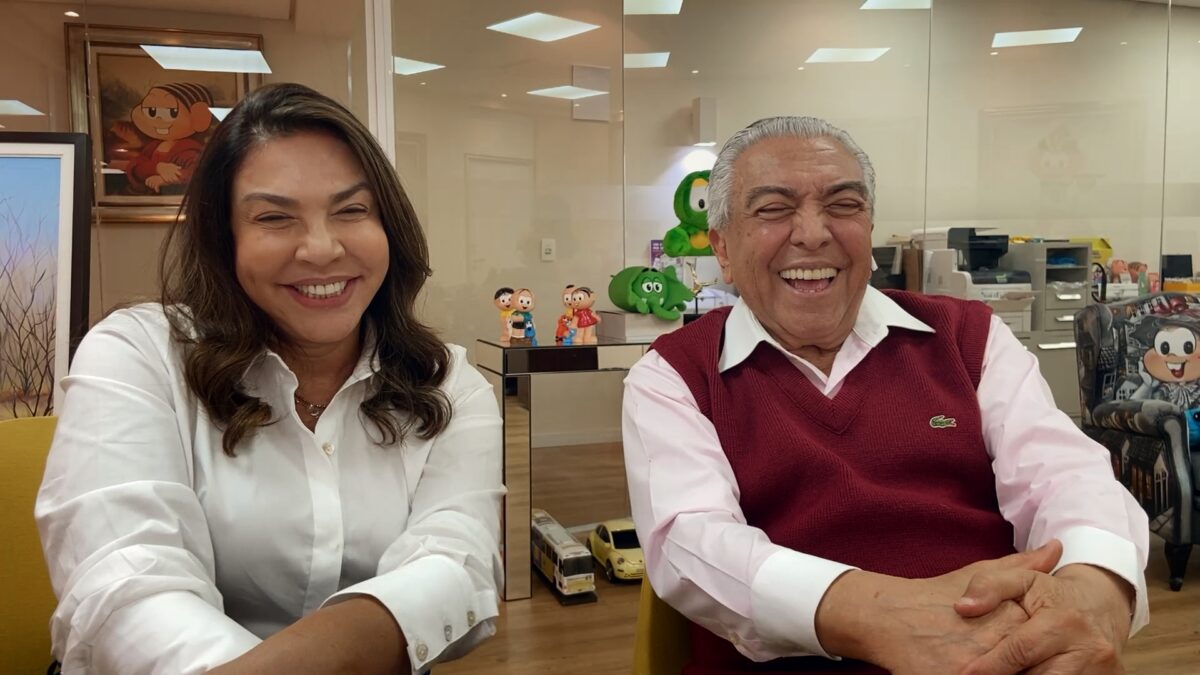 Mônica sorrindo, de camisa branca, sentada, de cabelo solto ao lado de Mauricio de Sousa, seu pai, de camisa social branca e sobreposição vinho, sorrindo durante gravação do Conversa com Bial, da TV Globo