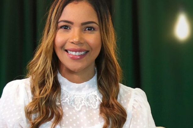 Cantora Amanda Wanessa de camisa manga longa branca, sorrindo, em estúdio com fundo verde escuro e uma lâmpada, durante ensaio fotográfico