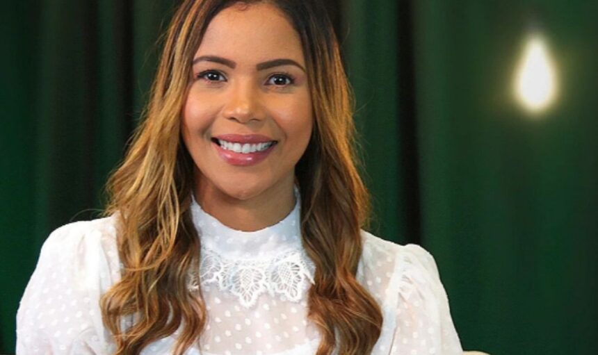 Cantora Amanda Wanessa de camisa manga longa branca, sorrindo, em estúdio com fundo verde escuro e uma lâmpada, durante ensaio fotográfico