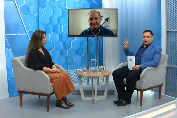 Eduardo Miranda sentado nos estúdios do Aparecida Debate, com um dos convidados sentado a sua frente e outro através de videoconferecia, pela televisão no centro do estúdio