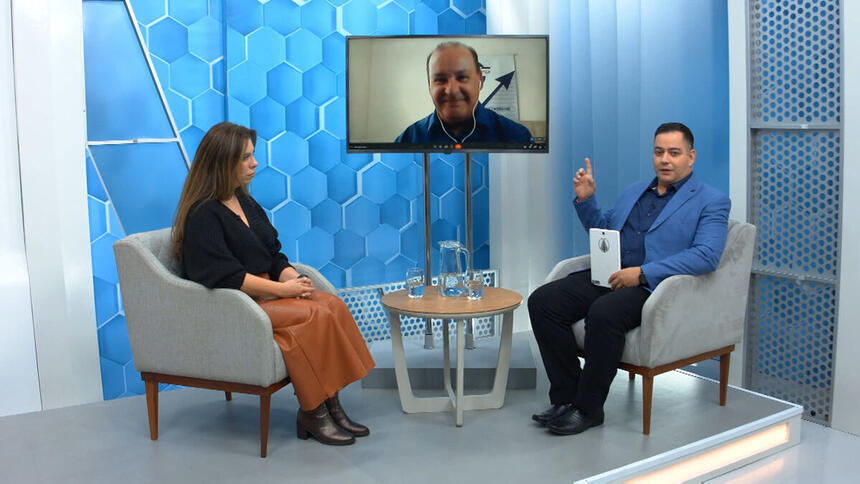 Eduardo Miranda sentado nos estúdios do Aparecida Debate, com um dos convidados sentado a sua frente e outro através de videoconferecia, pela televisão no centro do estúdio